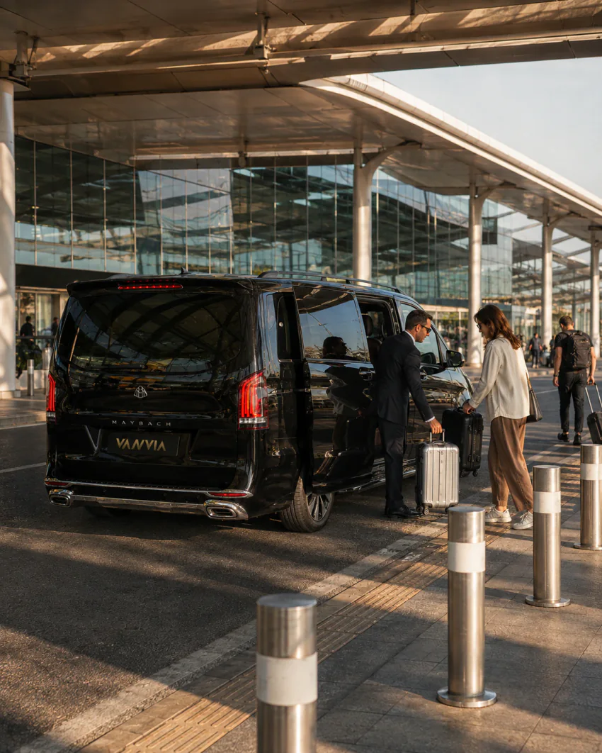 İzmir Havalimanı Akçay transferi için terminal saçağı altında valizli bir yolcuya yardım eden şoför ve siyah Vanvia aracının arka görünümü