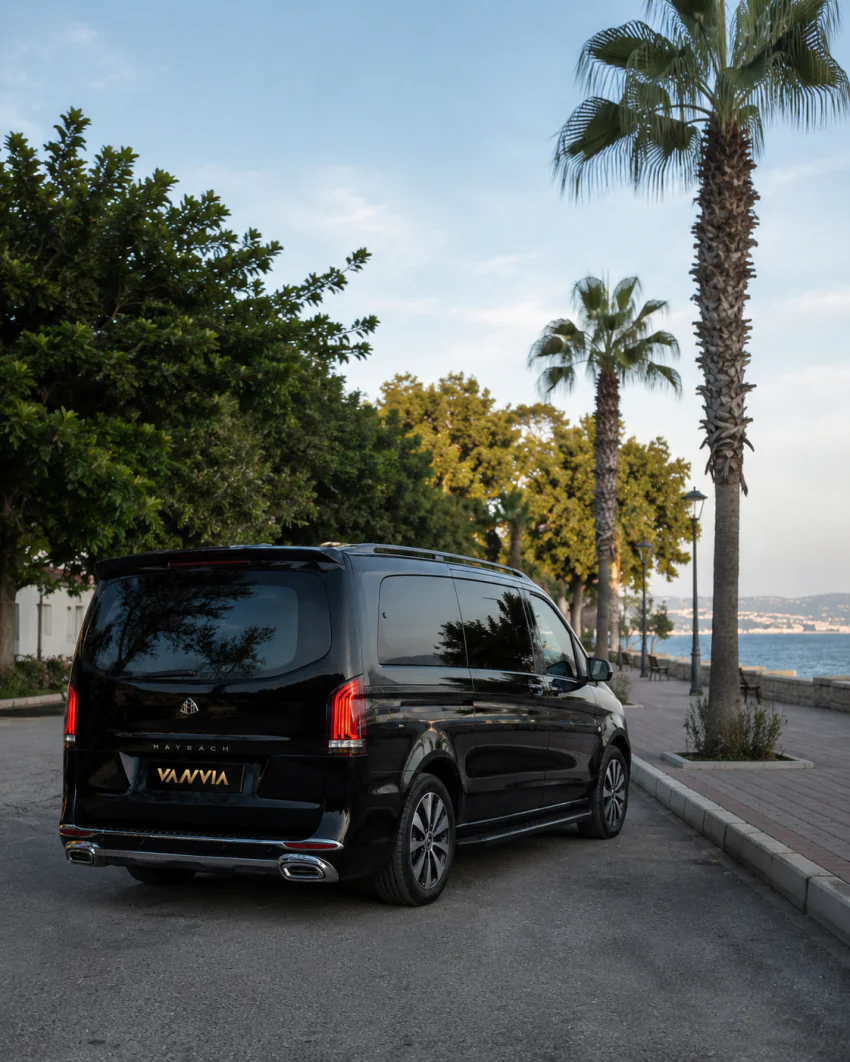 A rear view of a black Vanvia VIP transfer vehicle parked on a palm-lined coastal promenade at dusk, with the sea and a coastal town visible in the distance