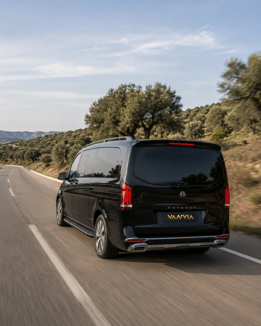 A rear view of a black Vanvia VIP transfer vehicle driving along an open road past a hillside covered in olive trees
