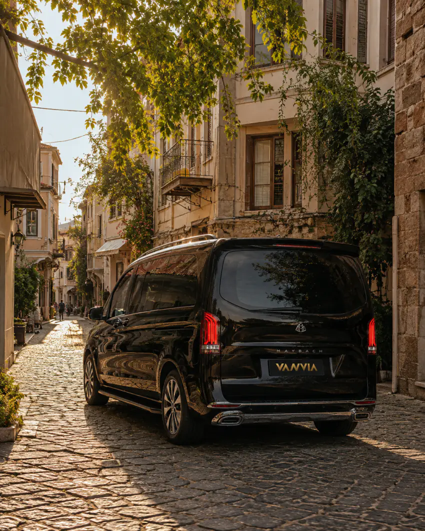 A rear view of a black Vanvia VIP transfer vehicle driving through a narrow cobblestone street lined with stone buildings at golden hour