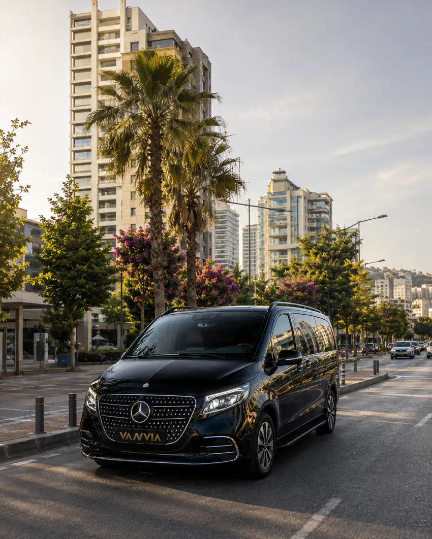 A black Vanvia VIP vehicle driving along a wide city boulevard lined with palm and flowering trees, with a tall building visible in the background