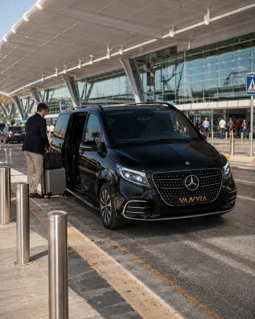 A front view of a black Vanvia VIP transfer vehicle waiting beside the driver loading luggage under the terminal canopy, for the Izmir Airport Burhaniye transfer