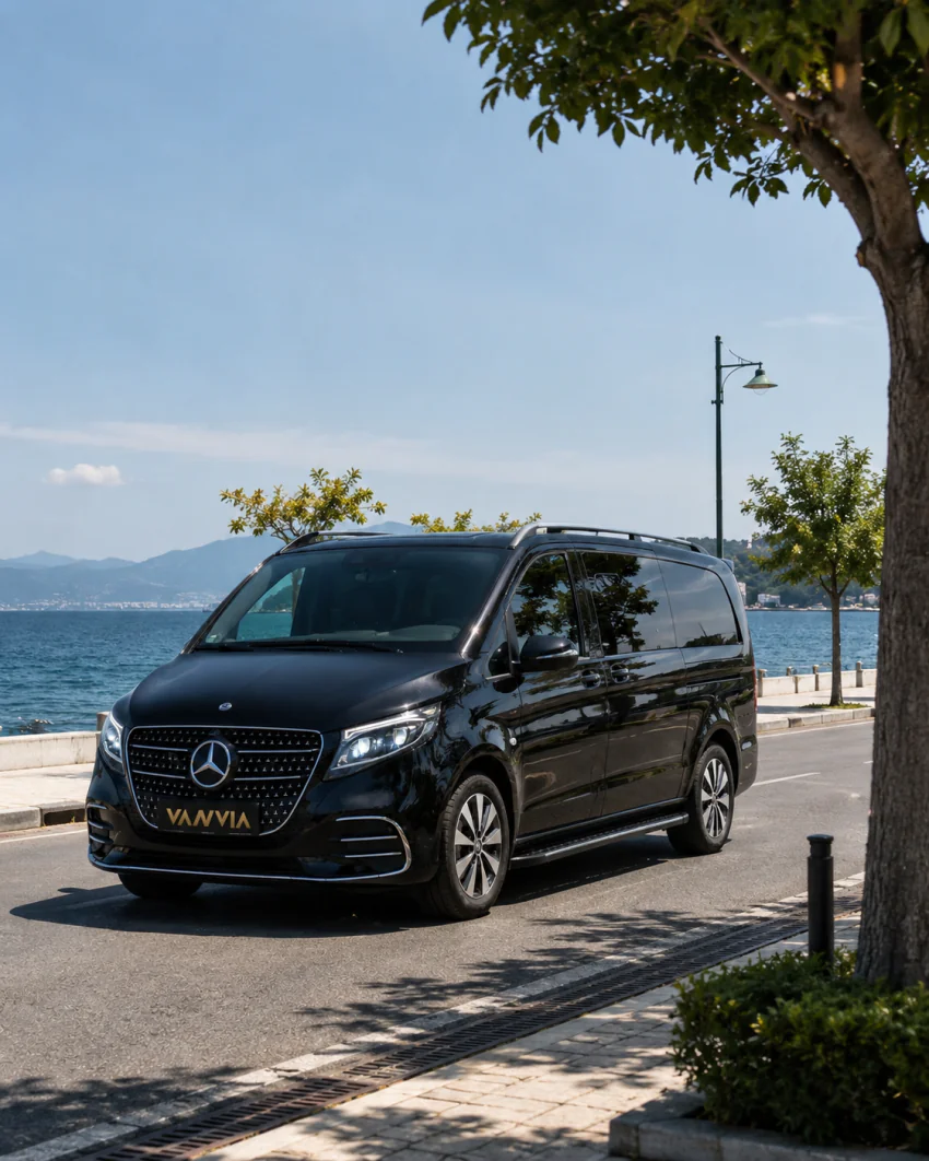 A black Vanvia VIP transfer vehicle parked on a waterfront road running alongside the sea, with hills visible across the water
