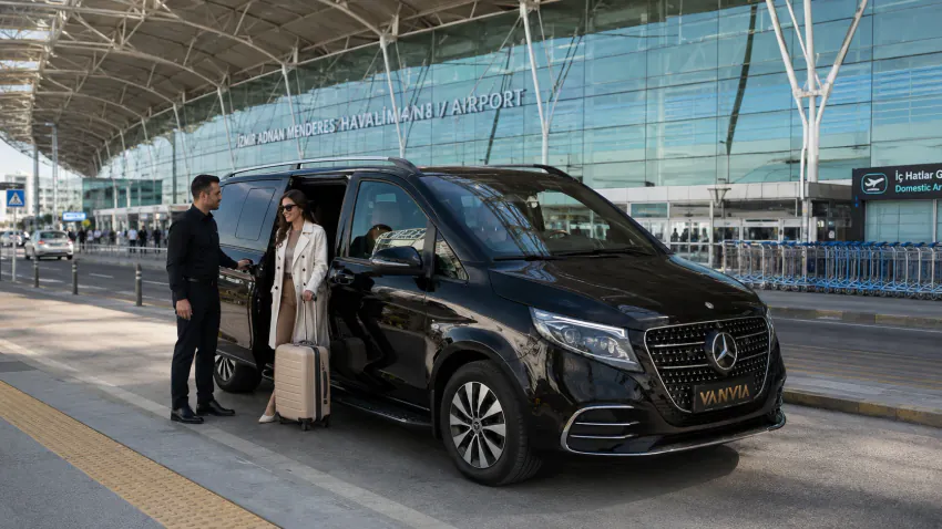 Rückansicht des Vanvia-VIP-Transferfahrzeugs vor dem Terminal des Flughafens Izmir Adnan Menderes, während der Fahrer die Tür für eine aussteigende Passagierin mit ihrem Koffer öffnet
