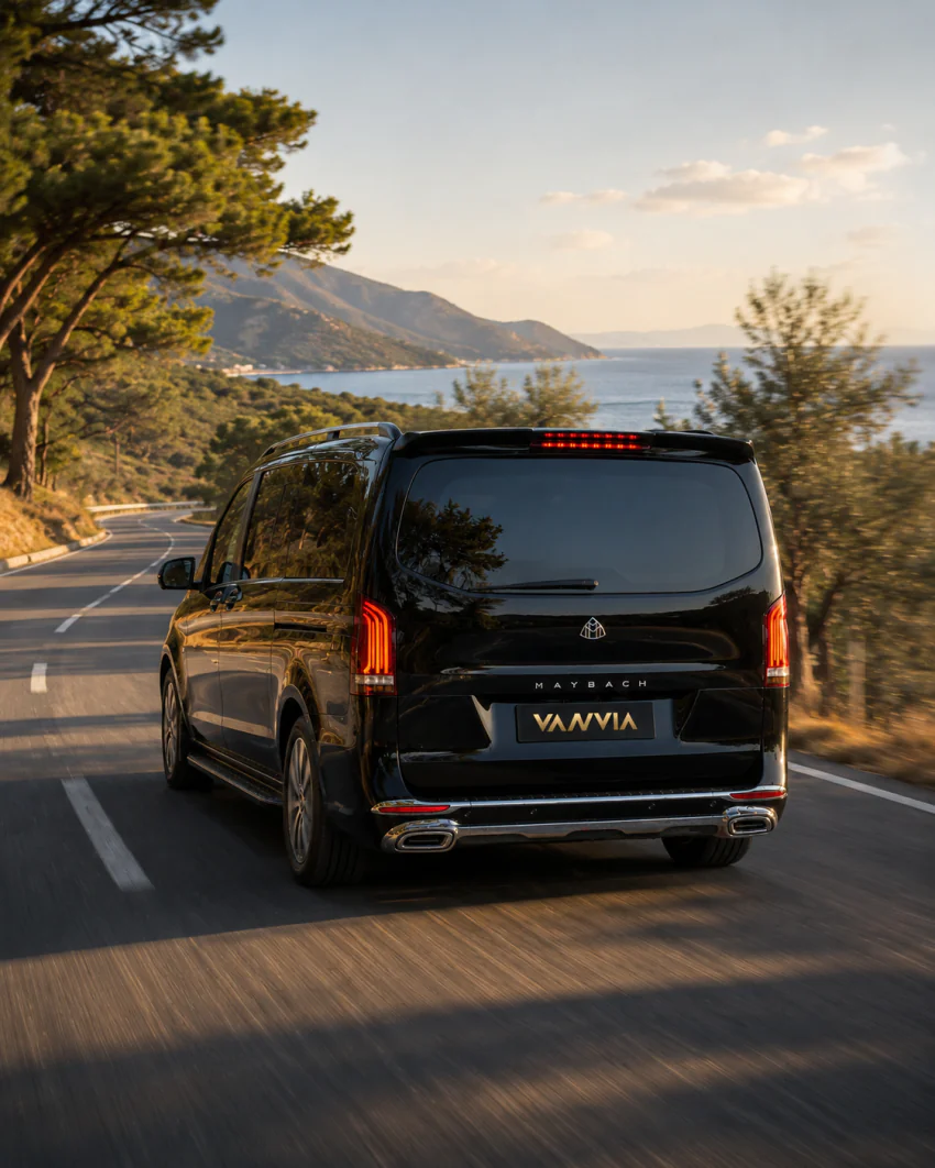 Black Vanvia VIP transfer vehicle driving along a pine-lined coastal road