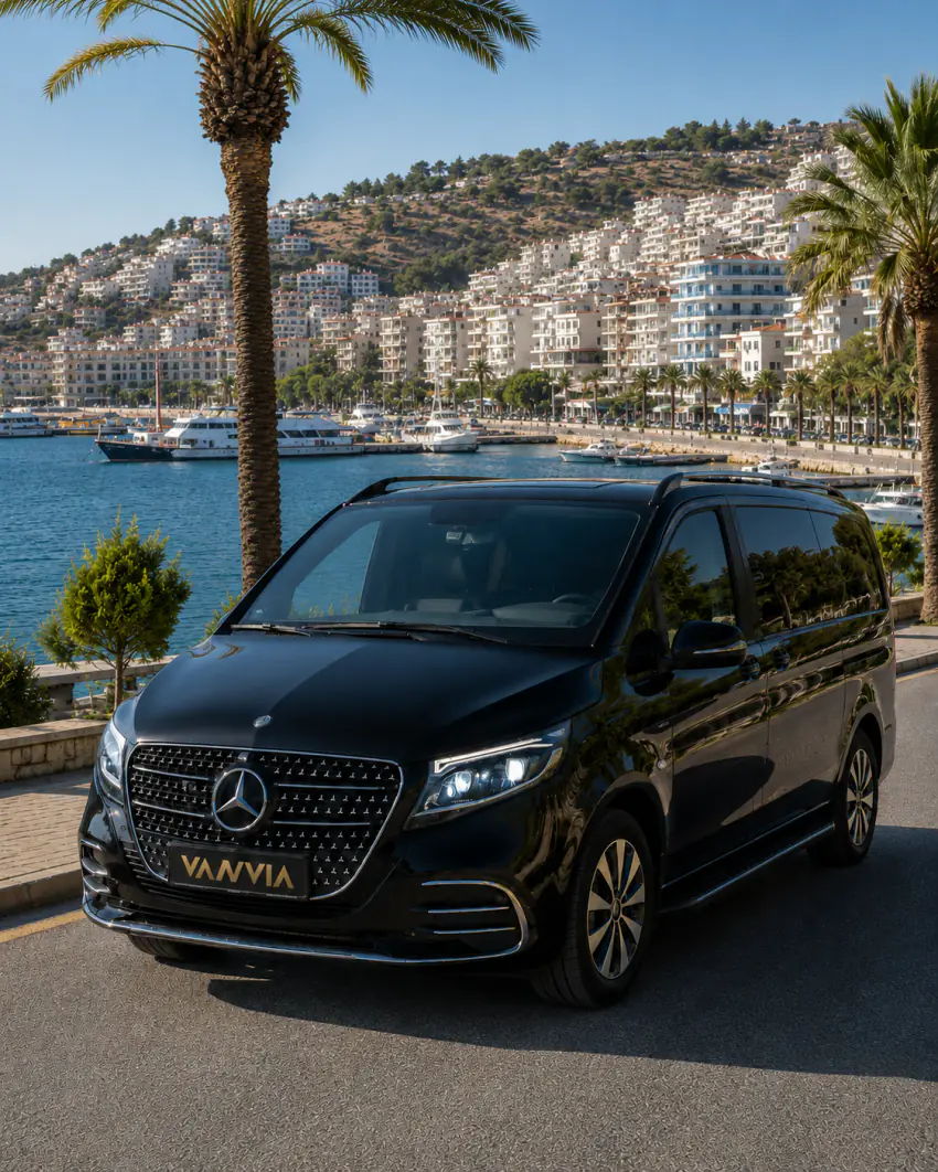 A black Vanvia VIP vehicle parked on a palm-lined coastal road, with a bay where numerous boats are moored and a dense hillside town visible in the background, for an Izmir Airport Kusadasi transfer, front view