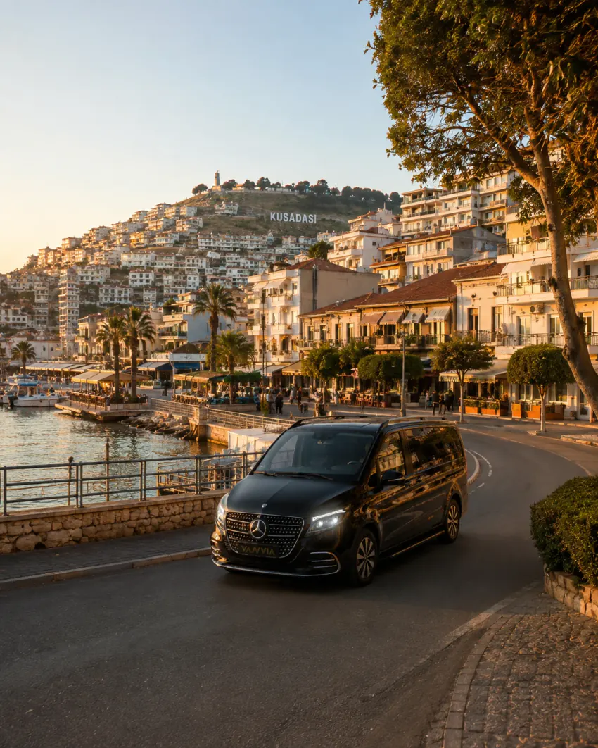 A black Vanvia VIP transfer vehicle driving along the coastal road at golden hour, with Kusadasi's hillside 'KUSADASI' sign and the waterfront town visible