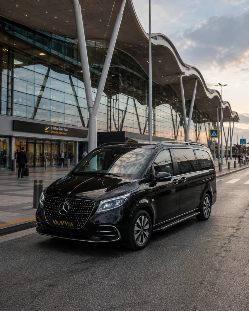 A black Vanvia VIP transfer vehicle at sunrise in front of the terminal building marked "İzmir Adnan Menderes Airport," with a passenger walking with luggage in the background
