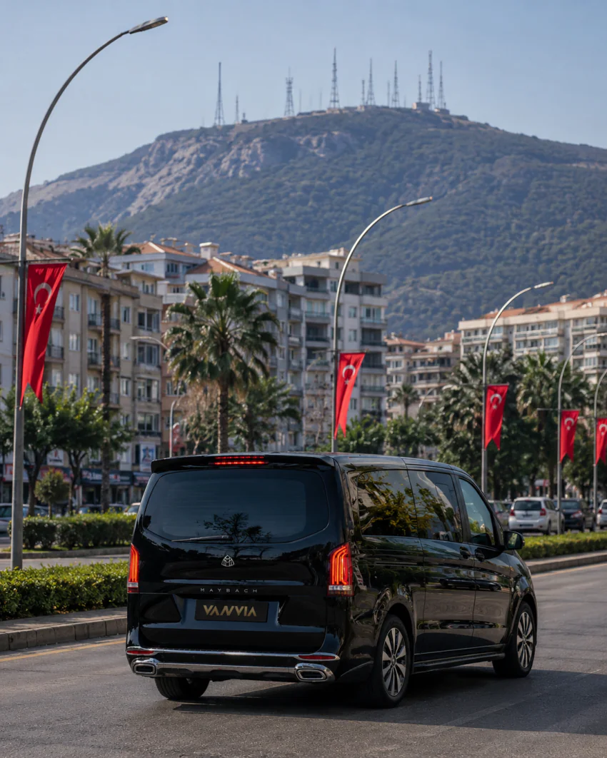 Vanvia Mercedes-Benz Vito VIP driving along a flag-lined boulevard on the Izmir Airport Manisa transfer route, with a mountain silhouette in the background