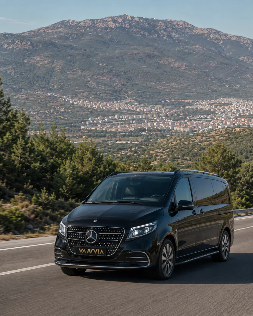 Vanvia Mercedes-Benz Vito VIP driving on a pine-lined mountain road toward Manisa, with a hillside settlement visible in the valley behind