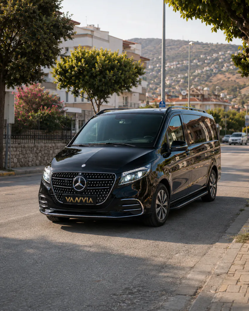 İzmir Havalimanı Menderes transfer için, arka planda tepelik bir yerleşim alanının göründüğü bir mahalle sokağında duran siyah Vanvia VIP aracının ön açıdan görünümü