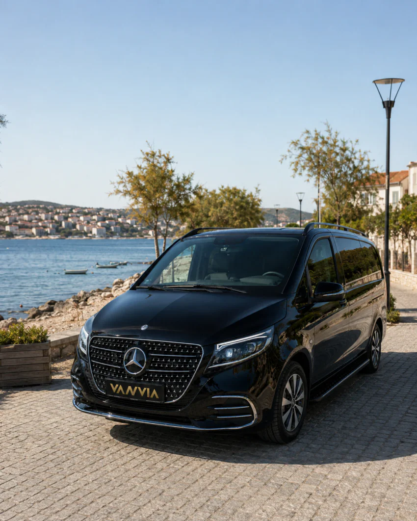A black Vanvia VIP vehicle driving along a road with a hillside coastal town visible in the background, for an Izmir Airport Seferihisar transfer