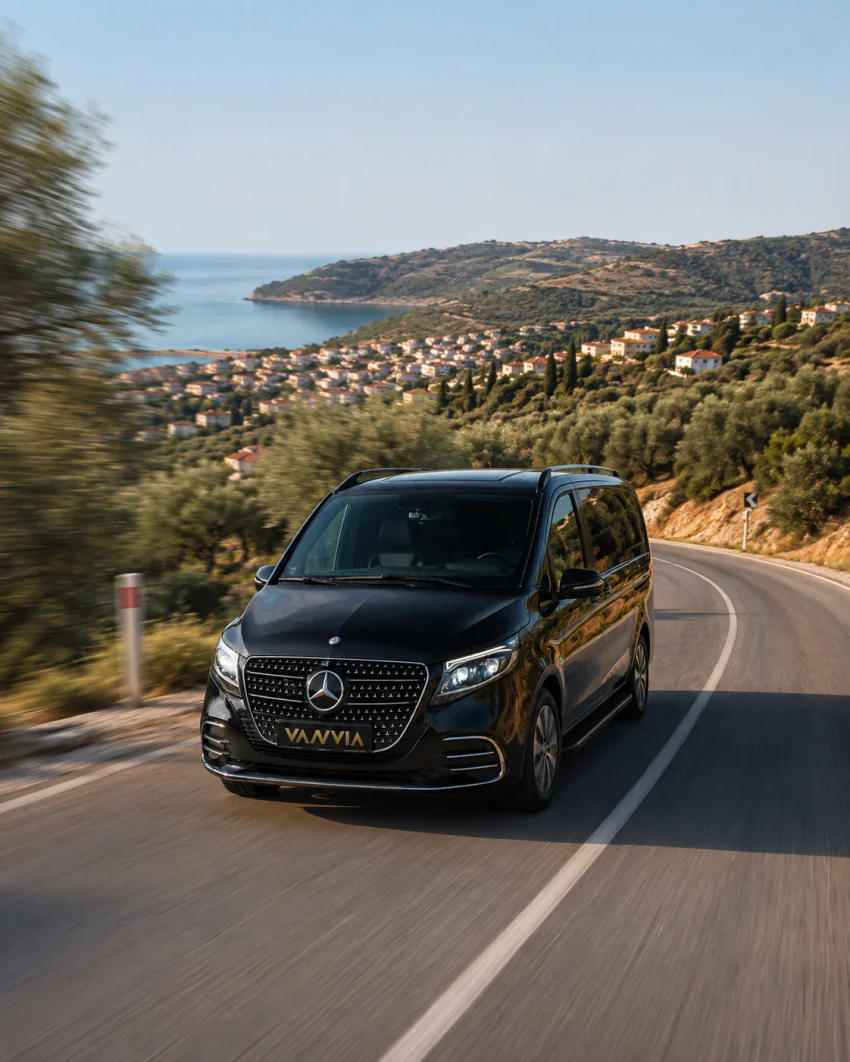 A black Vanvia VIP vehicle driving along a coastal road lined with olive trees, with a bay and a distant coastal settlement visible