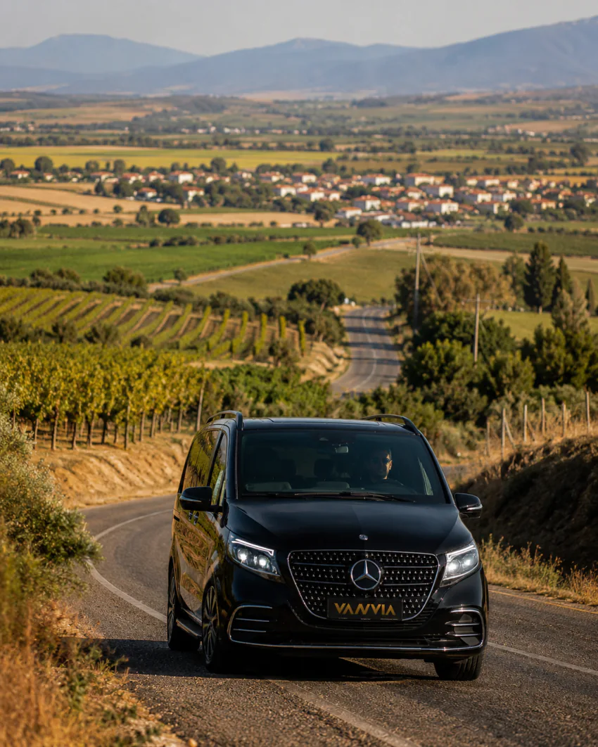 Vanvia Mercedes-Benz Vito VIP driving on a road lined with vineyards on both sides, with a red-roofed settlement visible in the valley below