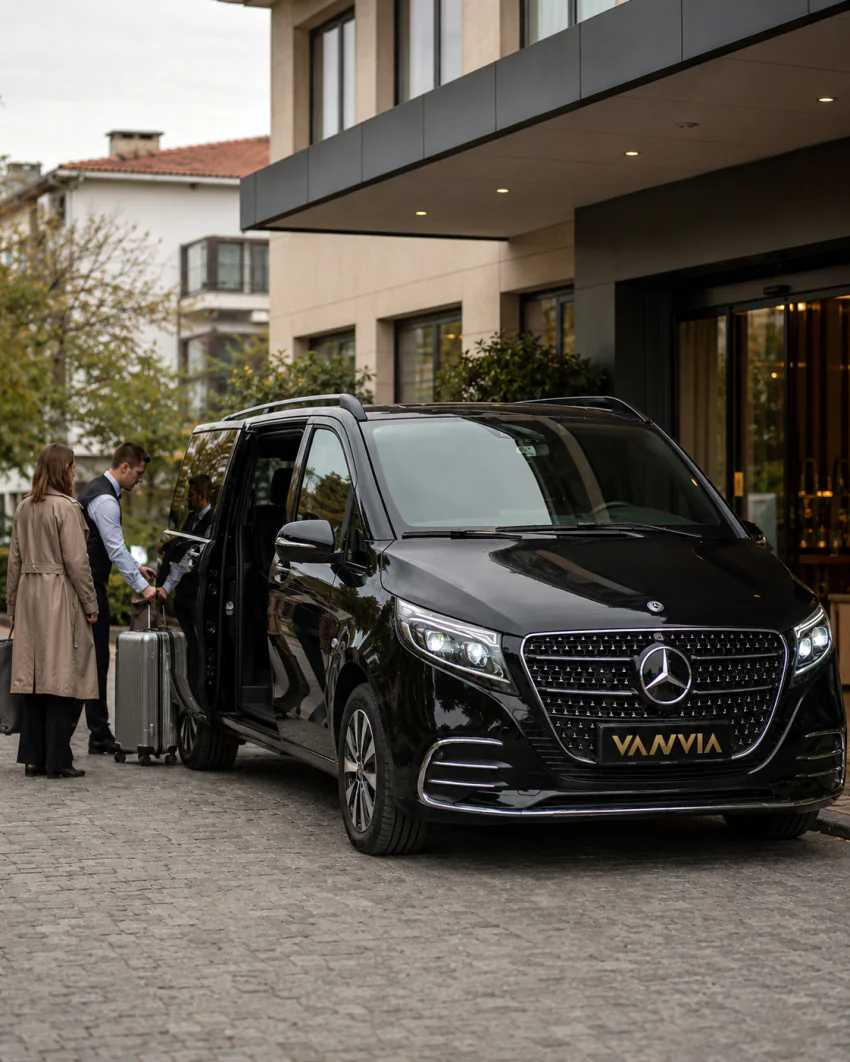 Vanvia chauffeur helping a traveler with a suitcase at a building entrance, with the vehicle door open and waiting