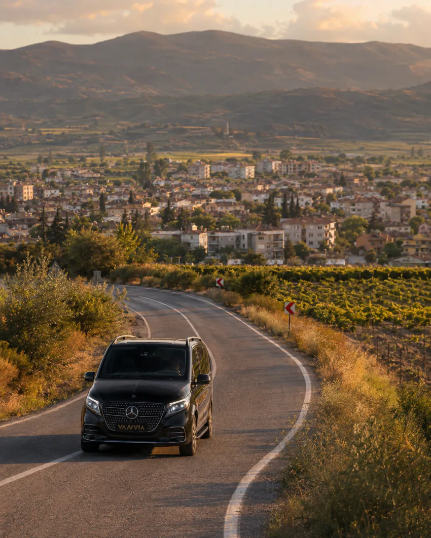 Vanvia Mercedes-Benz Vito VIP driving a winding road lined with farmland at sunset on the Izmir Airport Turgutlu transfer route, with a small town visible in the valley below