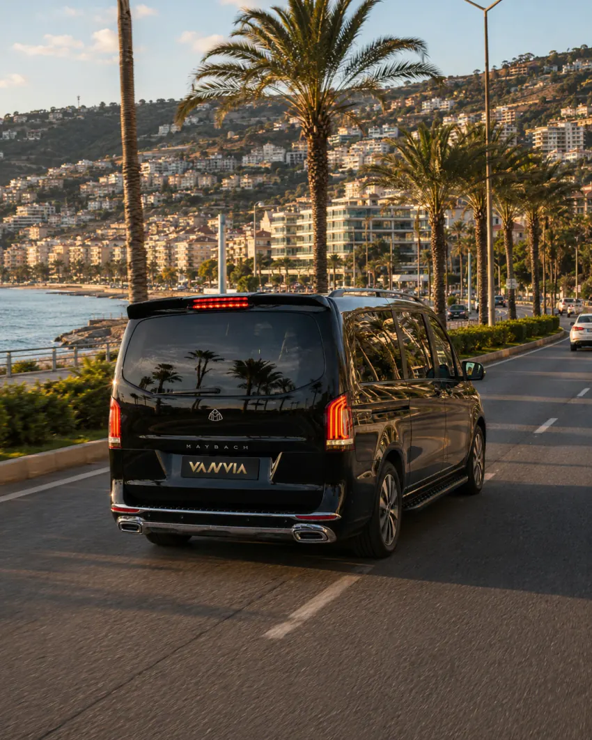 Alanya'da tepedeki apartman ve otel bölgesini arkasında bırakan sahil bulvarında altın ışıkta ilerleyen siyah Vanvia VIP transfer aracının arka üç çeyrek görünümü