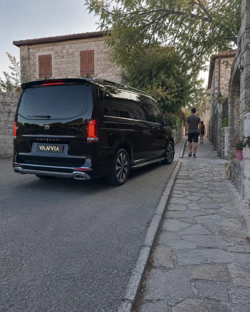 Heckansicht eines schwarzen Vanvia-VIP-Transferfahrzeugs, das auf einer schmalen, steingepflasterten Gasse in Behramkale hält; das Fahrzeug bleibt auf der befahrbaren Seite, während zwei Personen zu Fuß weitergehen