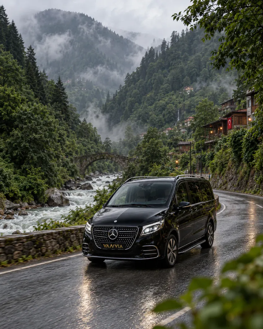A black Vanvia VIP transfer vehicle travels along a wet paved valley road beside a fast-running stream, with a stone arch bridge, wooden houses and mist-covered forested slopes behind