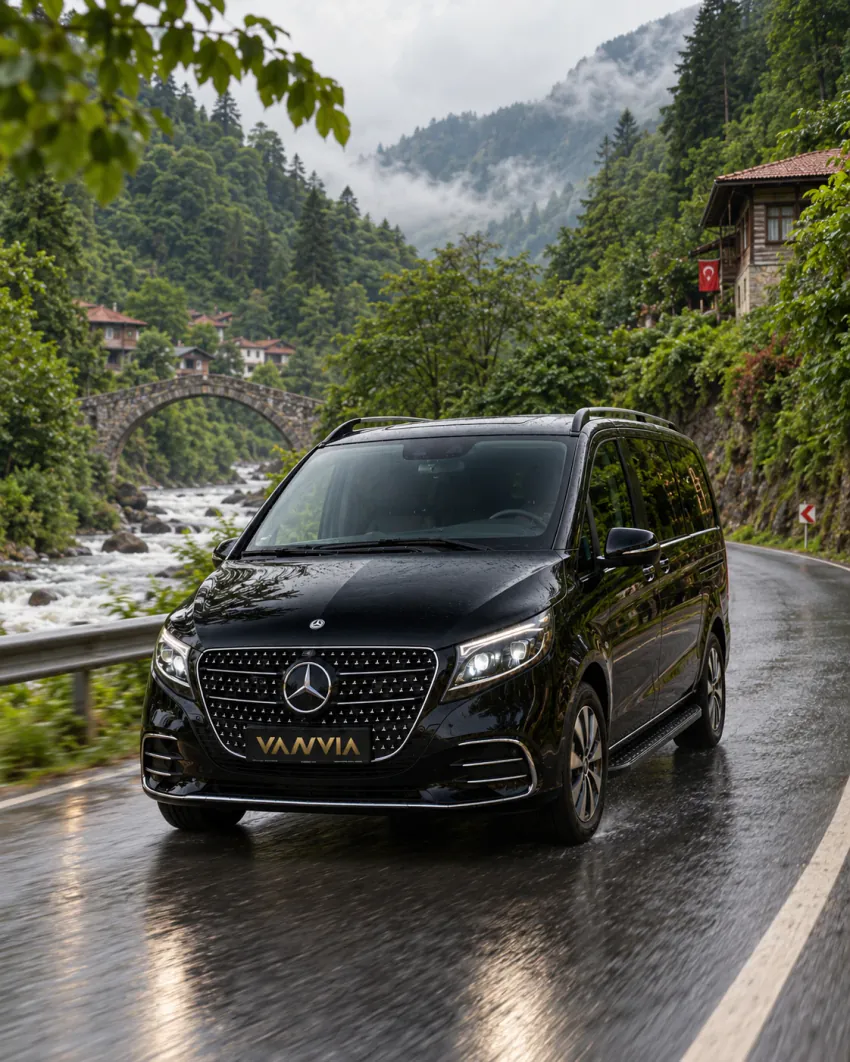 A black Vanvia VIP transfer vehicle on a wet two-lane public road with a guard rail on one side and a river on the other, village houses on the slope and misted forested mountains beyond