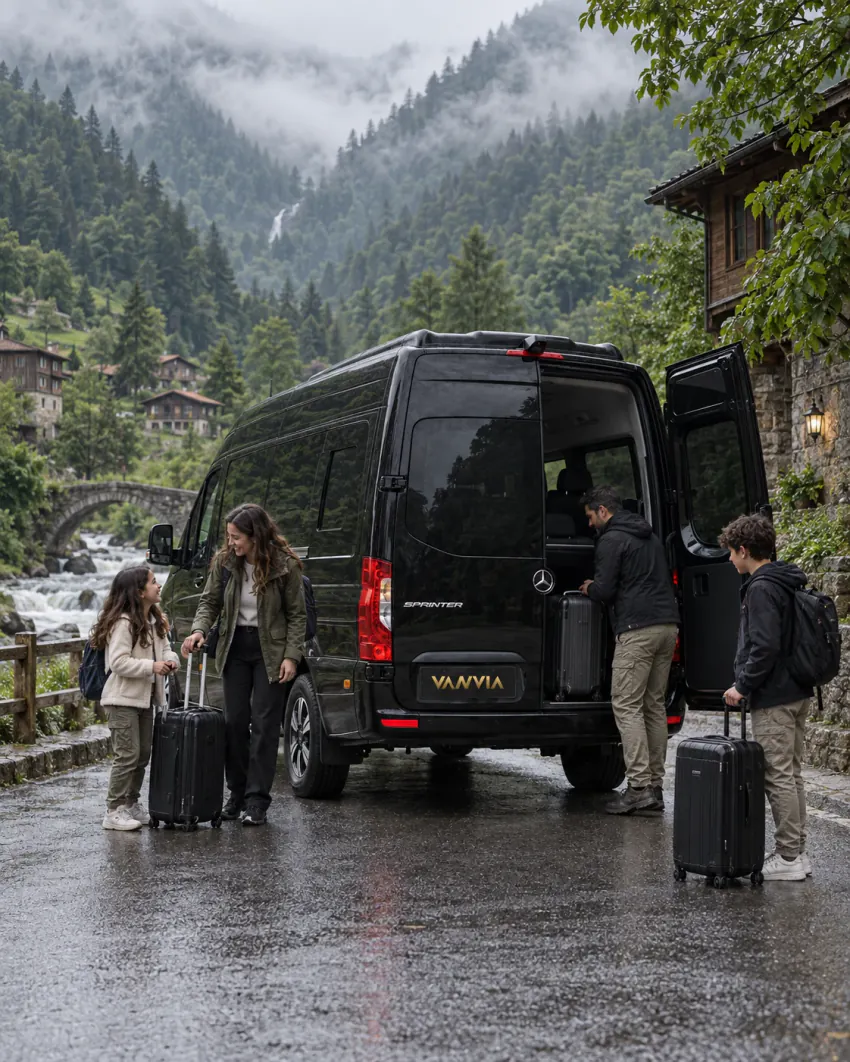 A black Vanvia VIP transfer van stopped at the edge of a wet public road with its side door open as a family boards with their own wheeled suitcases, wooden and stone buildings and misted mountains behind