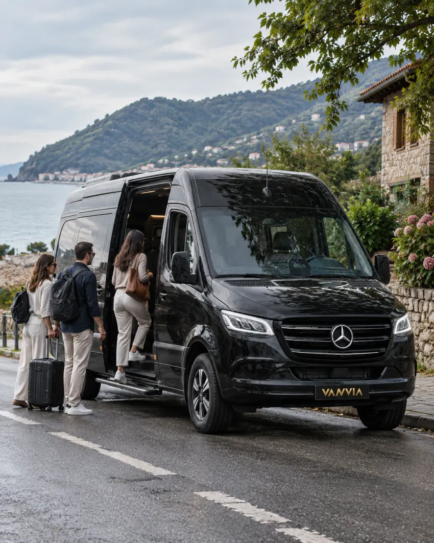 ركاب يصعدون بحقائبهم إلى حافلة صغيرة سوداء لخدمة Vanvia VIP transfer عبر بابها الجانبي المفتوح، على طريق عام قرب الساحل وخلفهم البحر وتلال حرجية ومبنى حجري