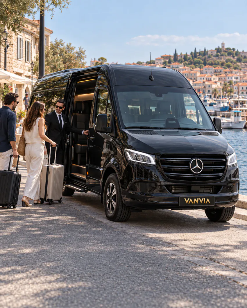 Passengers with their suitcases boarding a black Vanvia VIP transfer vehicle at a waterfront kerb lined with stone buildings and café umbrellas in Ayvalık, the driver waiting beside the open side door, with a hill of tiled-roof houses across the water