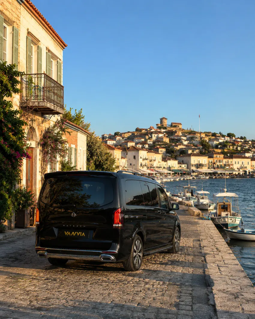 The rear view of a black Vanvia VIP transfer vehicle parked on the waterfront in Cunda (Alibey Island), beside a stone building, with moored fishing boats and a hillside settlement topped by a tower visible across the water