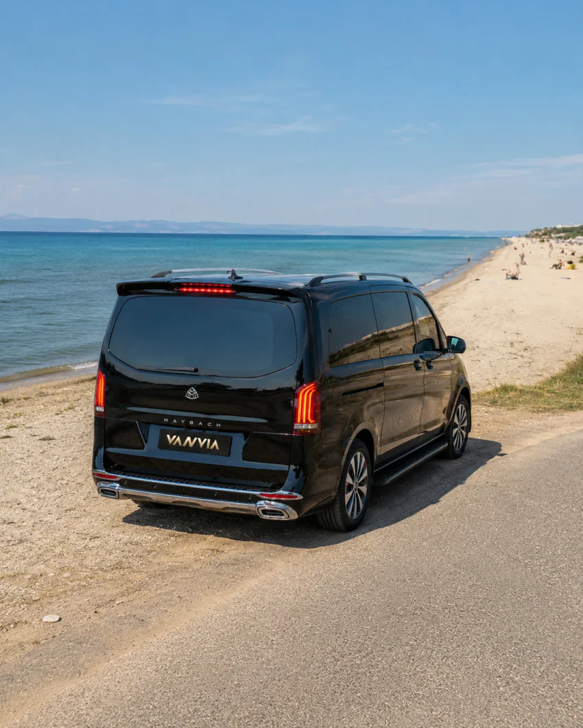 The rear view of a black Vanvia VIP transfer vehicle parked beside the road at Sarımsaklı beach, with the long sandy shoreline and turquoise sea visible, people on the beach in the distance