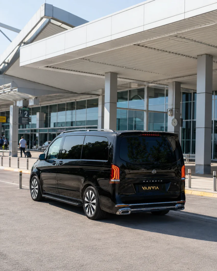A black Vanvia VIP transfer vehicle waiting at the kerb in front of the Milas-Bodrum Airport terminal building, with passengers nearby