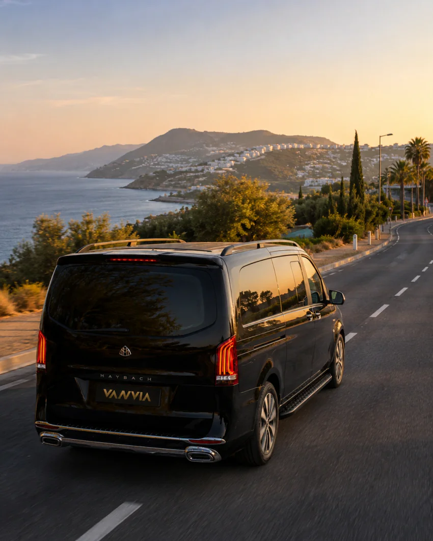 A rear view of a black Vanvia VIP transfer vehicle driving along the Bodrum peninsula's coastal road at sunset, with a white hillside town in the distance
