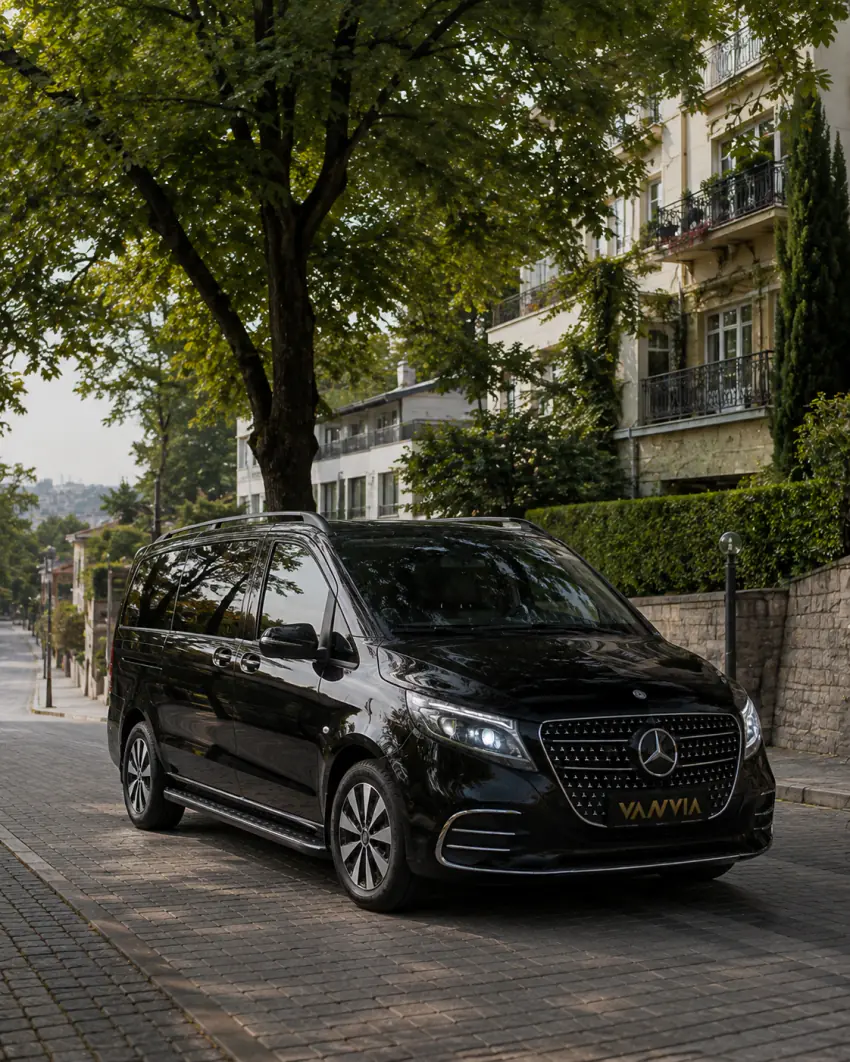 Frontansicht (Dreiviertelperspektive) eines schwarzen Vanvia-VIP-Transferfahrzeugs, das unter einem großen Baum auf einer Kopfsteinpflasterstraße vor einem efeubewachsenen Gebäude steht