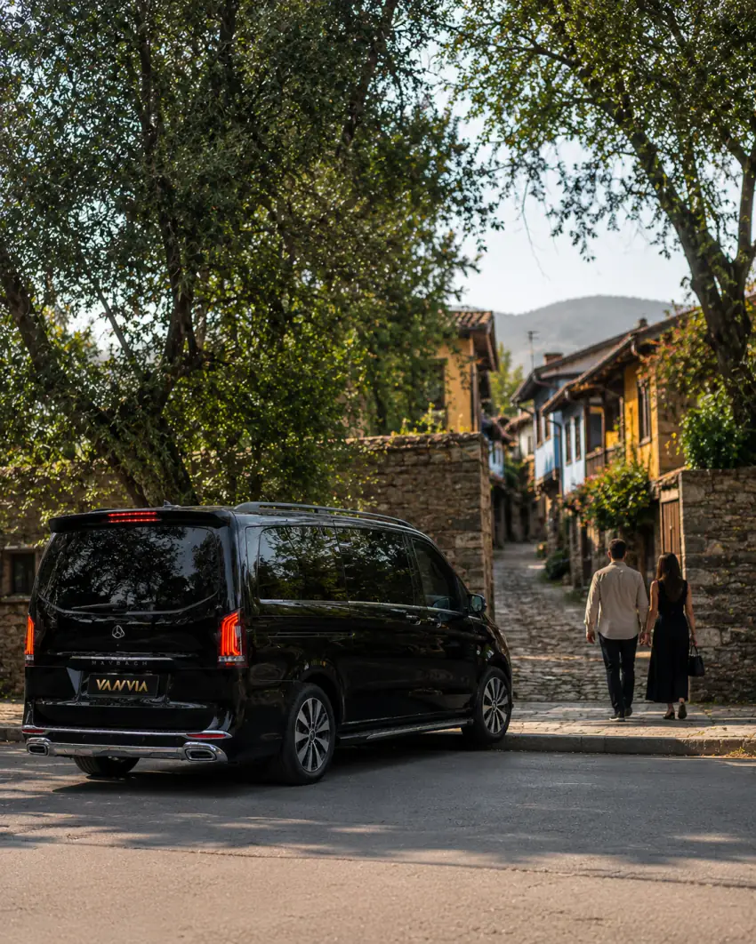 Heckansicht (Dreiviertelperspektive) eines schwarzen Vanvia-VIP-Transferfahrzeugs, das am straßenzugänglichen Rand einer schmalen, von historischen Stein- und Holzhäusern gesäumten Gasse steht, weiter vorne geht ein Paar auf dem gepflasterten Weg