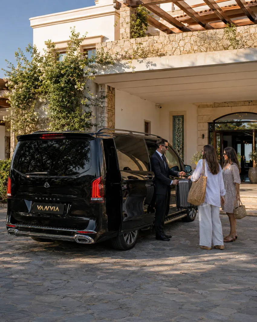 منظر خلفي جانبي لسيارة نقل VIP سوداء من فانفيا عند مدخل فيلا حجرية، والسائق يسلّم حقيبة للضيوف الواصلين تحت مظلة مغطاة بزهور الجهنمية