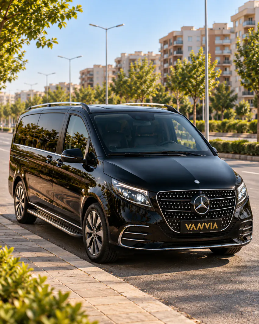 Ağaçlı geniş bir bulvarın kenarında park etmiş siyah Vanvia VIP transfer aracının ön çapraz görünümü; arkada apartman blokları var