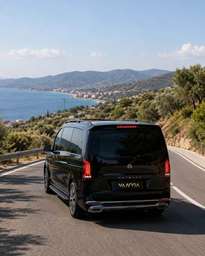 The rear view of a black Vanvia VIP transfer vehicle descending a mountain road lined with olive trees toward Ayvalık in daylight, with the sea and a coastal settlement visible in the distance