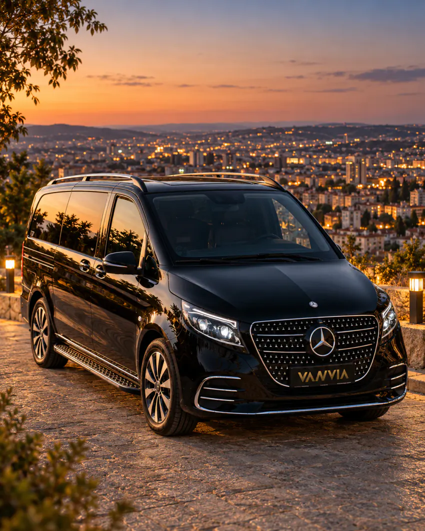 Akşam alacakaranlığında, ışıkları yanan şehrin genişçe görüldüğü yüksek bir seyir noktasında taş döşeli zeminde park etmiş siyah Vanvia VIP transfer aracı
