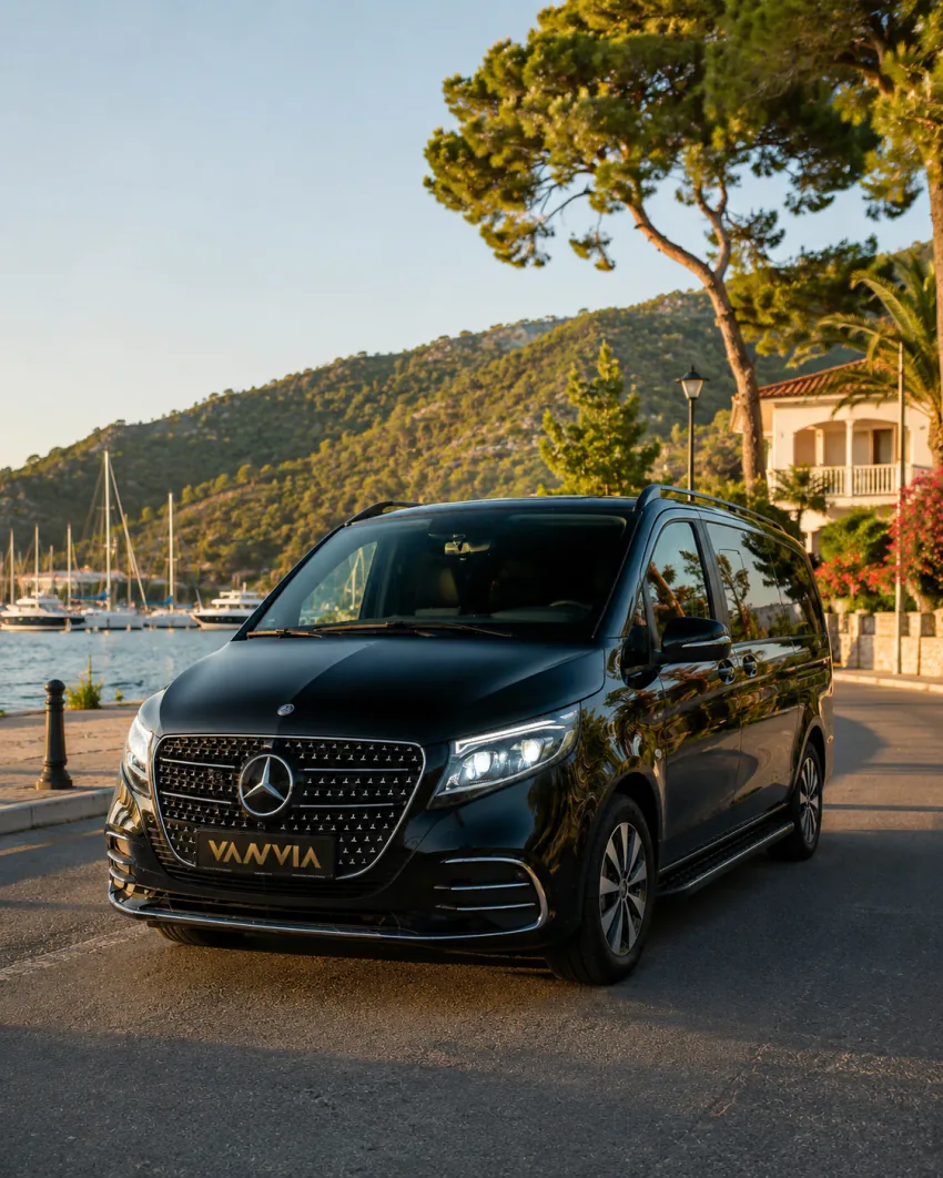 A front three-quarter view of a black Vanvia VIP transfer vehicle parked on the coastal road at Göcek bay by day among palm and pine trees, with yachts and the hillside town behind it