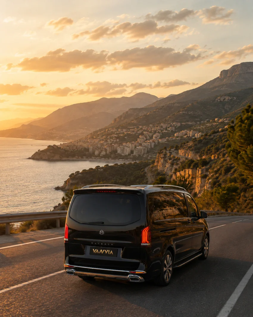 Alanya yönündeki dağlık kıyı yolunda, uzakta sahildeki bir yerleşim yerinin göründüğü bir seyir noktasında gün batımında duran siyah Vanvia VIP transfer aracının arka görünümü, koruma bariyeri önde