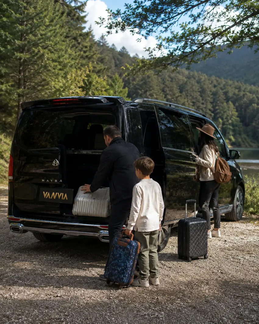 Eine Familie verstaut Koffer im offenen Kofferraum eines schwarzen Vanvia-VIP-Transferfahrzeugs, im Hintergrund Wald und See