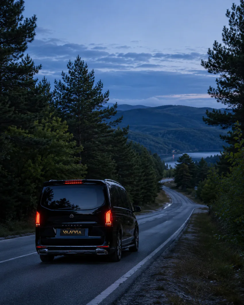 Rückansicht eines schwarzen Vanvia-VIP-Transferfahrzeugs, das in der Dämmerung eine Waldstraße entlangfährt, in der Ferne ein See und Berge