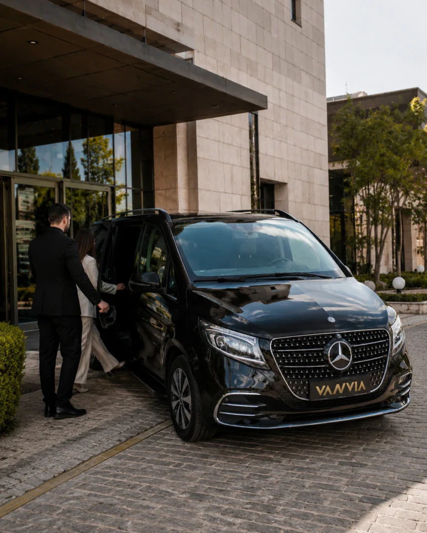 Ein schwarzes Vanvia-VIP-Transferfahrzeug am Eingang eines modernen Hotels, der Fahrer hält die Tür, während Gäste hineingehen