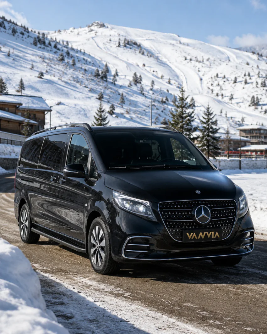 Frontansicht eines schwarzen Vanvia-VIP-Transferfahrzeugs, das auf einer schneebedeckten Bergstraße steht, dahinter Skichalets und ein schneebedeckter Gipfel