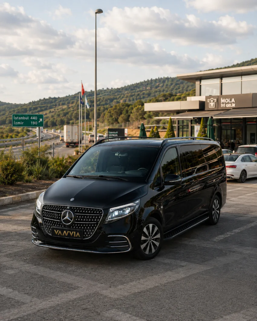 Schräge Frontansicht eines schwarzen Vanvia-VIP-Transferfahrzeugs an einer Autobahnraststätte, in der Nähe ein Entfernungsschild Istanbul–İzmir