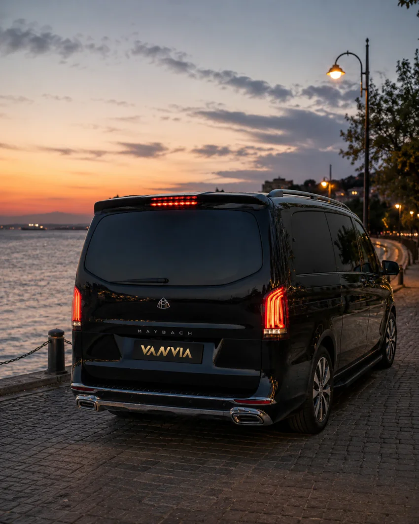 Rückansicht eines schwarzen Vanvia-VIP-Transferfahrzeugs, das bei Dämmerung an einer Küstenpromenade neben dem Meer und Straßenlaternen steht