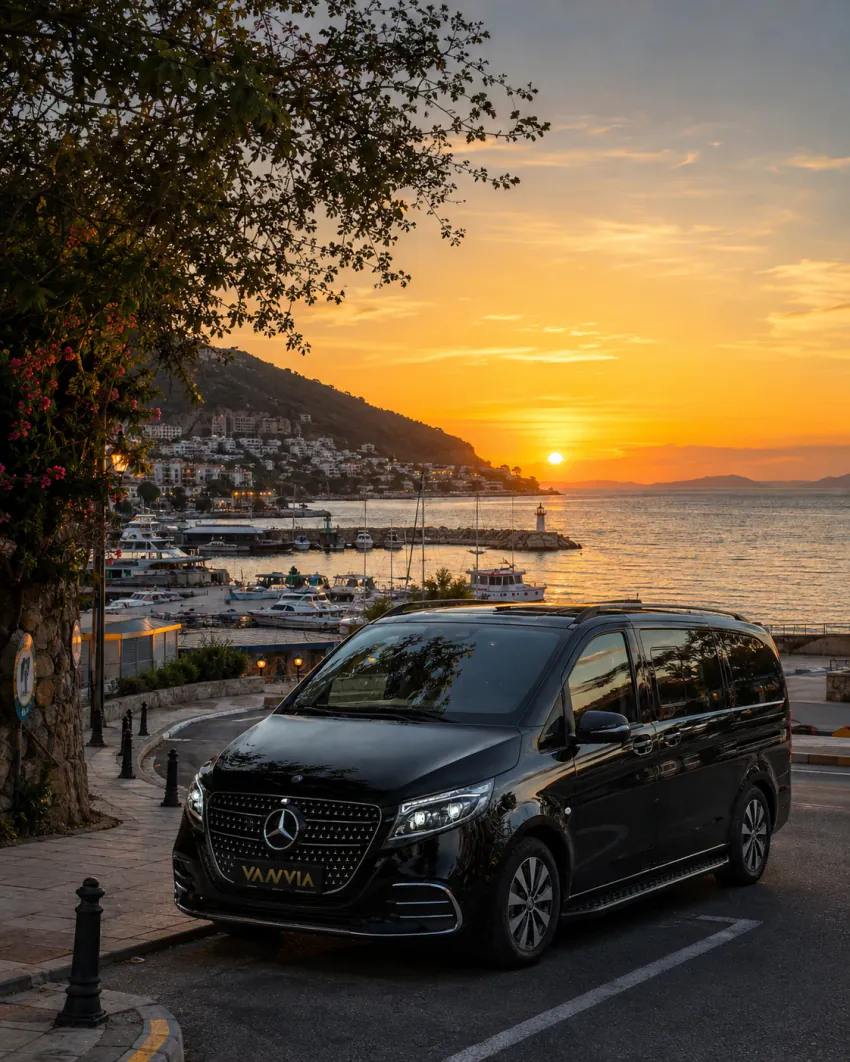 Kaş'ta fenerli küçük bir liman ve demirli teknelerin göründüğü bir yol kenarında gün batımında duran siyah Vanvia VIP transfer aracının ön görünümü, arkada yamaçtaki kasaba