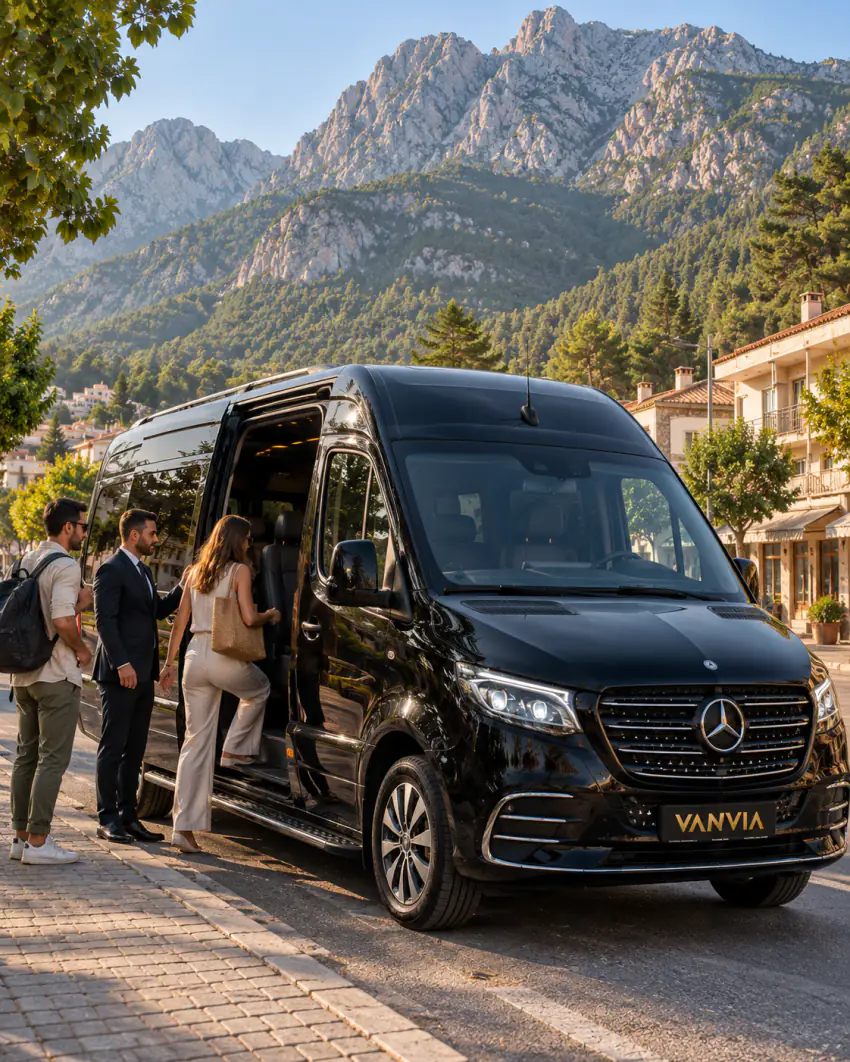 Passengers boarding a black Vanvia VIP transfer vehicle at an ordinary town-street kerb in Kemer, with the jagged Taurus Mountains and pine-covered slopes rising behind, the driver waiting beside the open side door