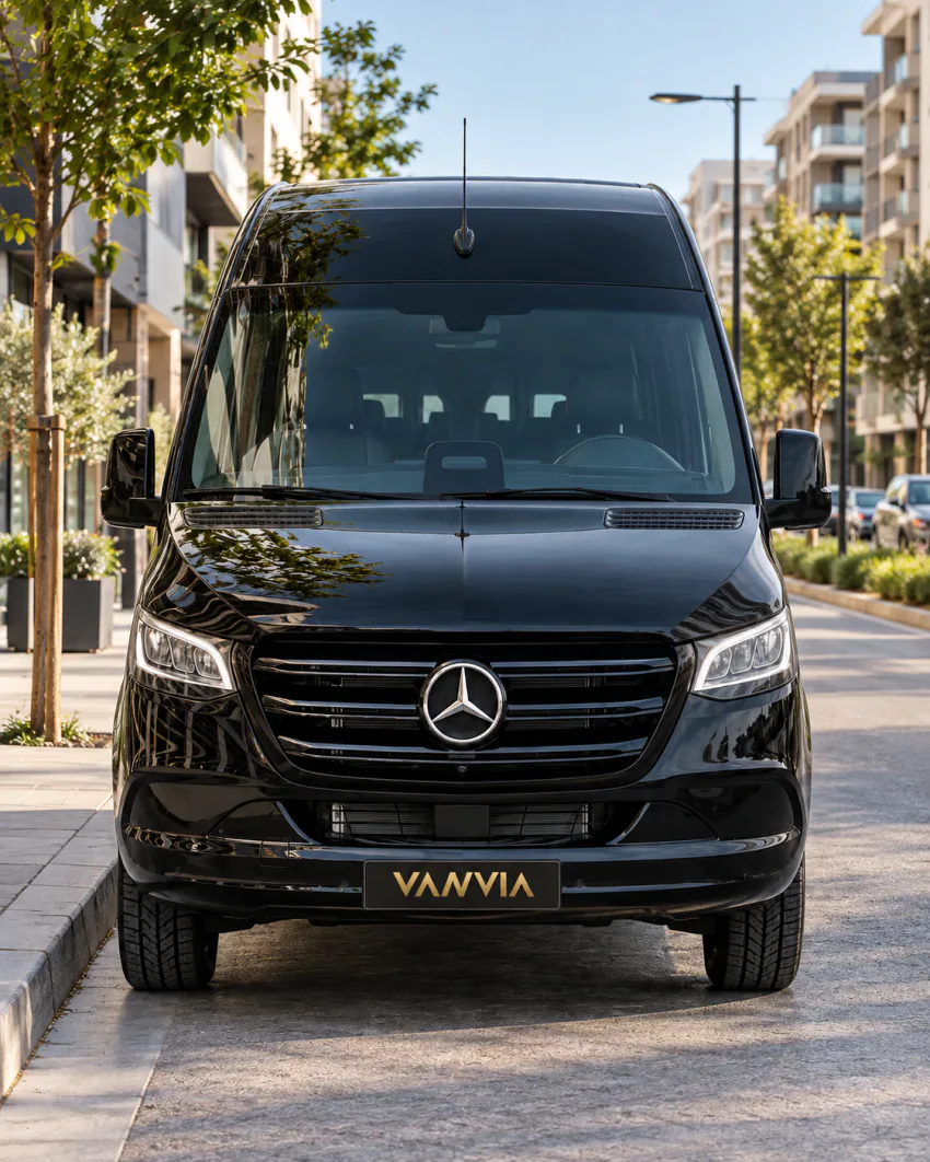 Genç ağaçlar ve modern apartmanlarla çevrili bir şehir caddesinde kaldırım kenarında duran siyah Vanvia VIP transfer aracının önden görünümü