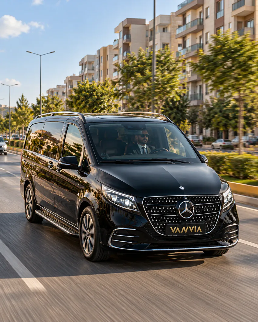 Mardin VIP transfer: ağaçlı ve açık renkli apartmanlarla çevrili geniş bir caddede ilerleyen siyah Vanvia aracı; direksiyonda takım elbiseli, güneş gözlüklü şoför görünüyor