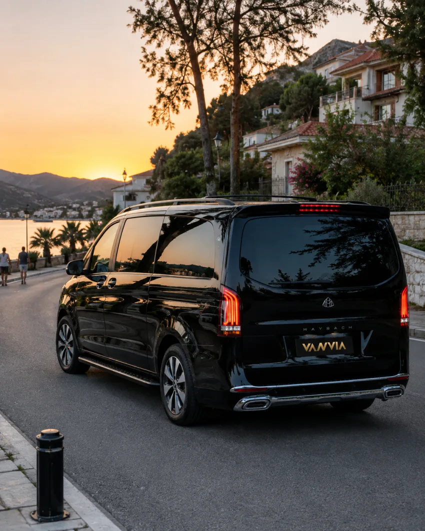 A rear view of a black Vanvia VIP transfer vehicle parked on the coastal road overlooking Marmaris's hillside town at sunset, with pedestrians on the promenade